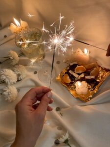 From above of unrecognizable female with burning Bengal light standing near flowers wineglass and food on tray on white fabric in room with burning candles