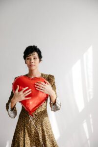 Serious female with red heart shaped balloon for Saint Valentine day standing against white wall